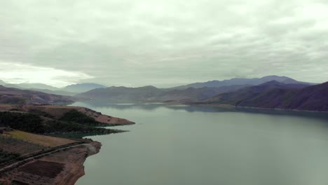 Fabulous-lake-surrounded-by-mountains-and-hills-with-agricultural-parcels,-fruit-trees-planted-on-shore,-aerial-shot