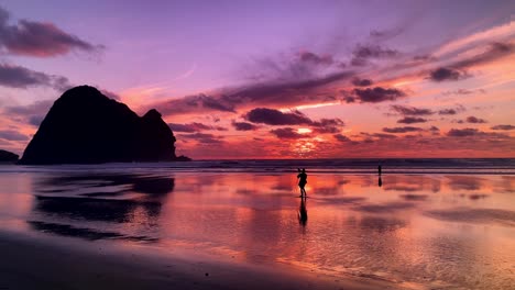 A-silhouetted-man-takes-a-photo-with-his-camera-and-tripod-on-a-beach-reflecting-a-beautiful,-multicolored-sunset-on-Piha-Beach-in-New-Zealand