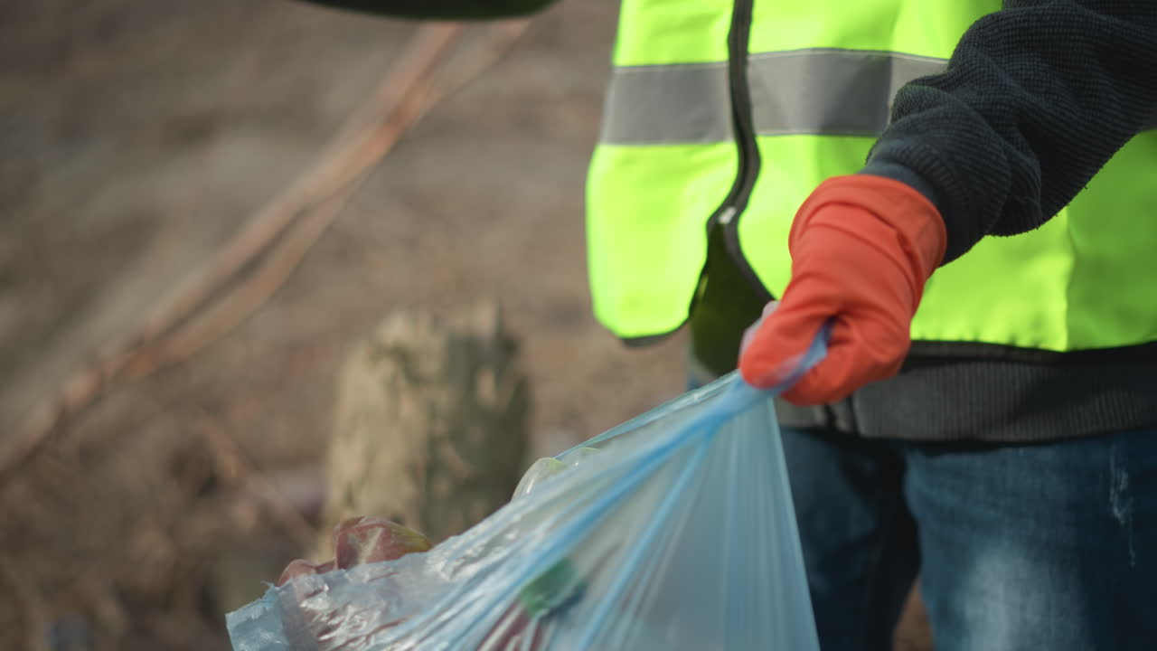 Volunteer wearing high visibility vest and black gloves reaching into pile of dry leaves and branches to pick up hidden litter during outdoor cleanup, removing waste to protect environment