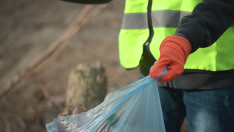 volunteer wearing high visibility vest and black gloves reaching into pile of dry leaves and branches to pick up hidden litter during outdoor cleanup, removing waste to protect environment