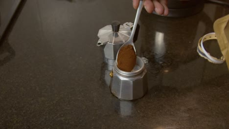 unidentified hand holding a spoon with ground coffee beans putting in the moka pot, preparing to brew coffee-closeup shot