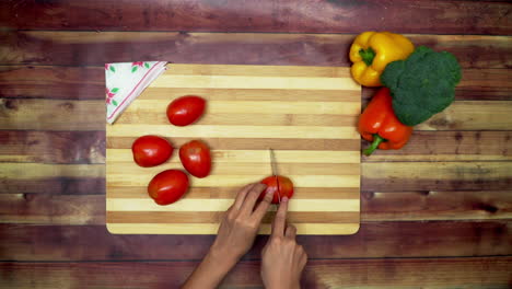 a top view of slicing the tomatoes by hand, two big yellow and red capsicums and a green broccoli on the table