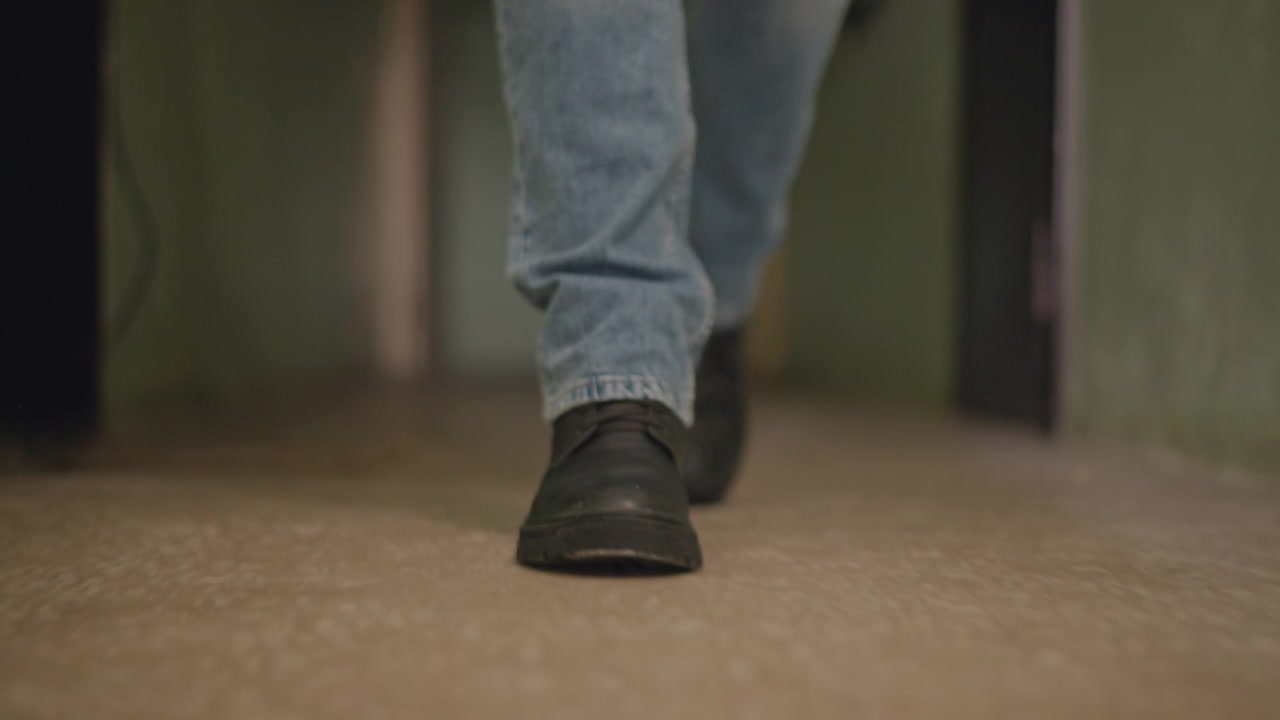 Ground-level view of adult moving through narrow indoor corridor, feet and lower legs in focus, subtle motion blur, steady stride, concrete floor, soft warm light, everyday urban commute