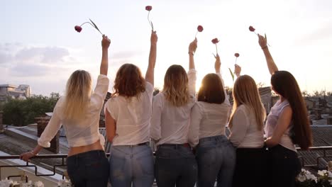 six seductive young women are standing on a terrace in a row from their back. wearing casual clothes, white shirts and jeans. raising up their hands with a flower. front view