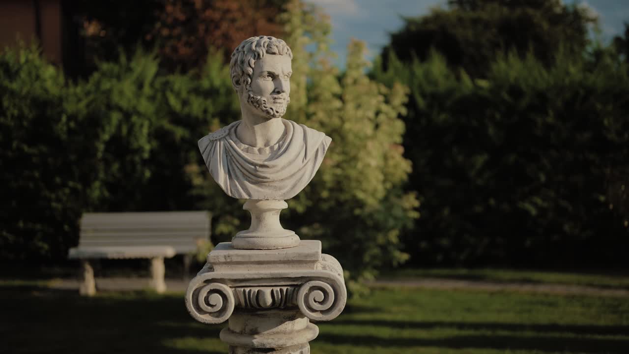 Bust of a classical male figure placed on a pedestal in a garden. The marble-style sculpture, with detailed beard and draped garment, stands against lush greenery