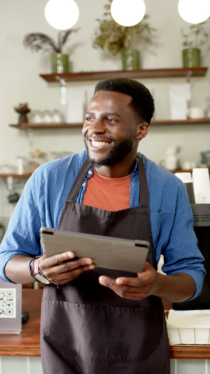 Vertical video: African American male barista holding tablet, smiling in cozy cafe
