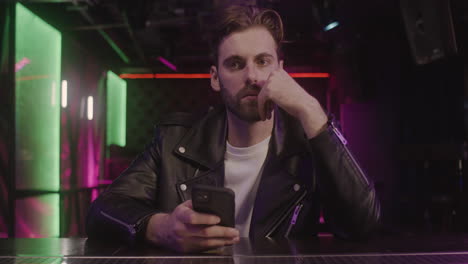 hombre barbudo serio usando teléfono móvil mientras está sentado en el mostrador del bar