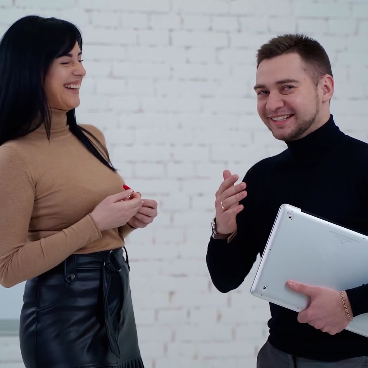 Smiling coworkers talking indoors. Handsome man holding a laptop and telling something to a young woman standing near the board for writing.