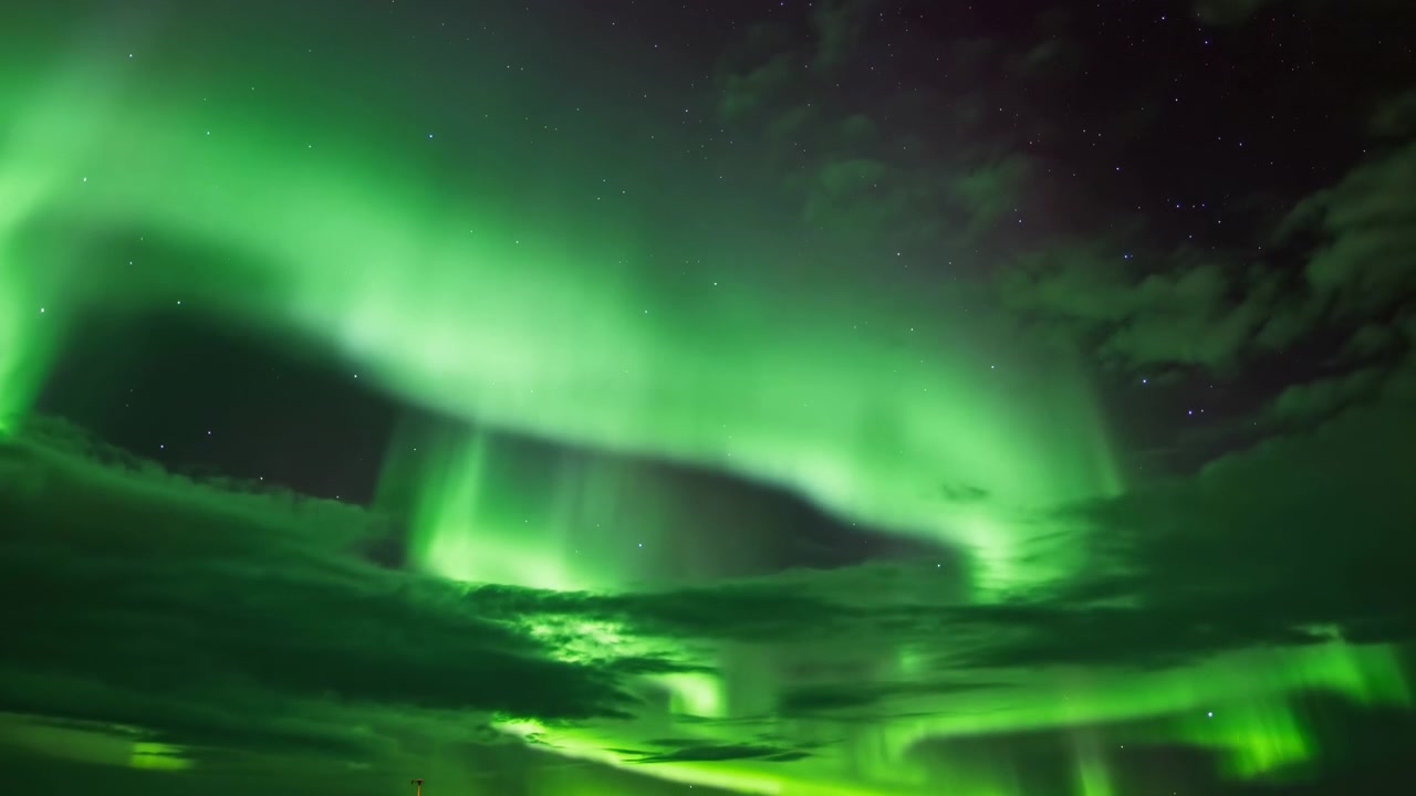 Un'aurora verde brillante riempie l'ampio cielo notturno, formando ampie forme a tenda e pilastri di luce verticali sopra nuvole stratificate, con un orizzonte luminoso, profondità scura e un'atmosfera polare tranquilla