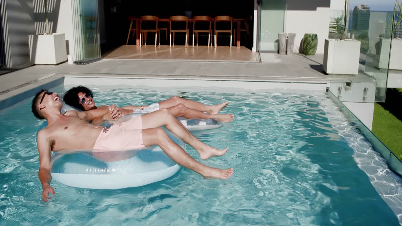 Sun urging male friends lounging, laughing on round float at private pool wearing trunks and shades