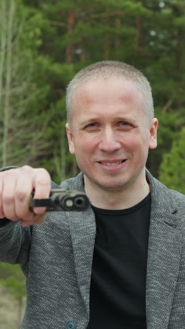 회색 재과 검은 셔츠를 입은 남자가 초록색 숲의 배경 앞에 서서 권총을 겨누고 있는 근접한 모습.