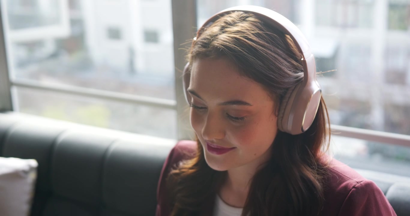 Adult woman in maroon blazer, pink headset sitting on tufted couch by window, tilting head, smiling