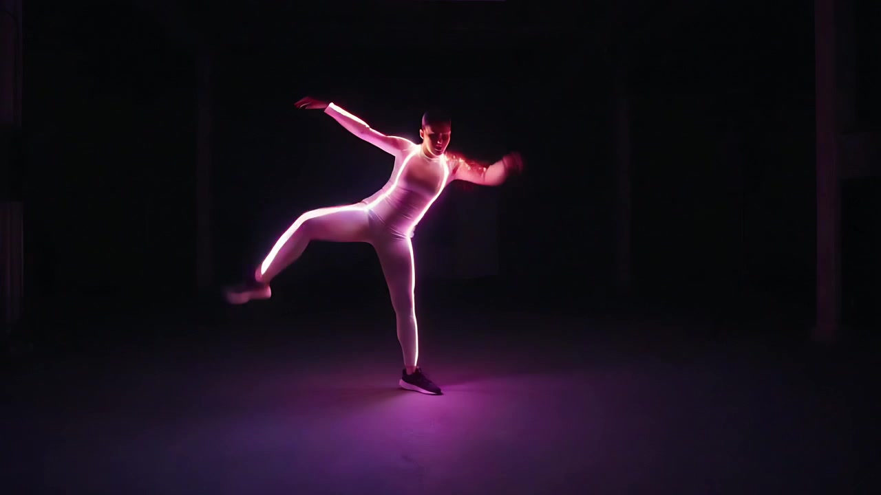 Donna che balla seguendo il cambio di colore dei LED, facendo passi, calci e mantenendo l'equilibrio in un magazzino, con la tuta luminosa.