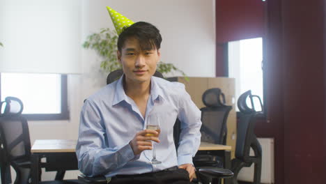 portrait of a confident man in formal outfit and party hat holding a champagne glass and looking at camera while sitting at the office