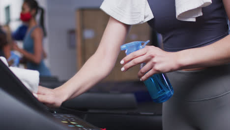 fit caucasian woman wearing face mask cleaning treadmill machine with disinfectant in the gym