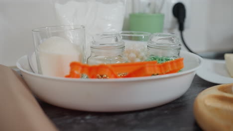 close-up of sugar in glass alongside small glass jars filled with colorful beads, baking tools, including orange cookie cutters, placed in a white bowl on a sleek black countertop