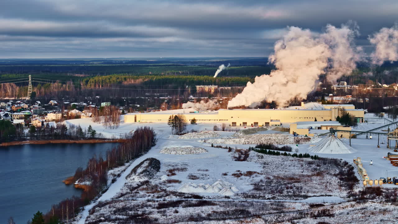 Dronebillede af et snedækket industriområde ved en sø, der udleder en tyk dampsky under vinterlige skyer, som viser energiproduktion i koldt vejr og miljøpåvirkning