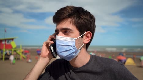 Joven-Respondiendo-Una-Llamada-Telefónica-En-La-Playa-Mientras-Usa-Una-Máscara-Debido-A-Una-Pandemia