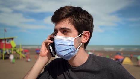 joven respondiendo una llamada telefónica en la playa mientras usa una mascarilla debido a una pandemia