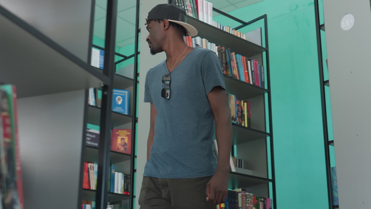 Young man in casual shirt and backward cap walking through modern library, glancing at bookshelves filled with colorful books, thoughtful expression