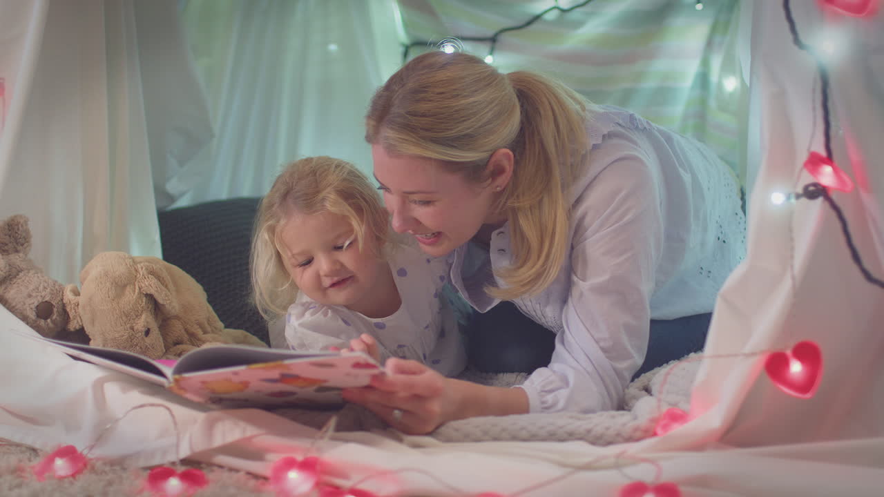 Mother and young daughter reading story in homemade camp in child's bedroom at home - shot in slow motion