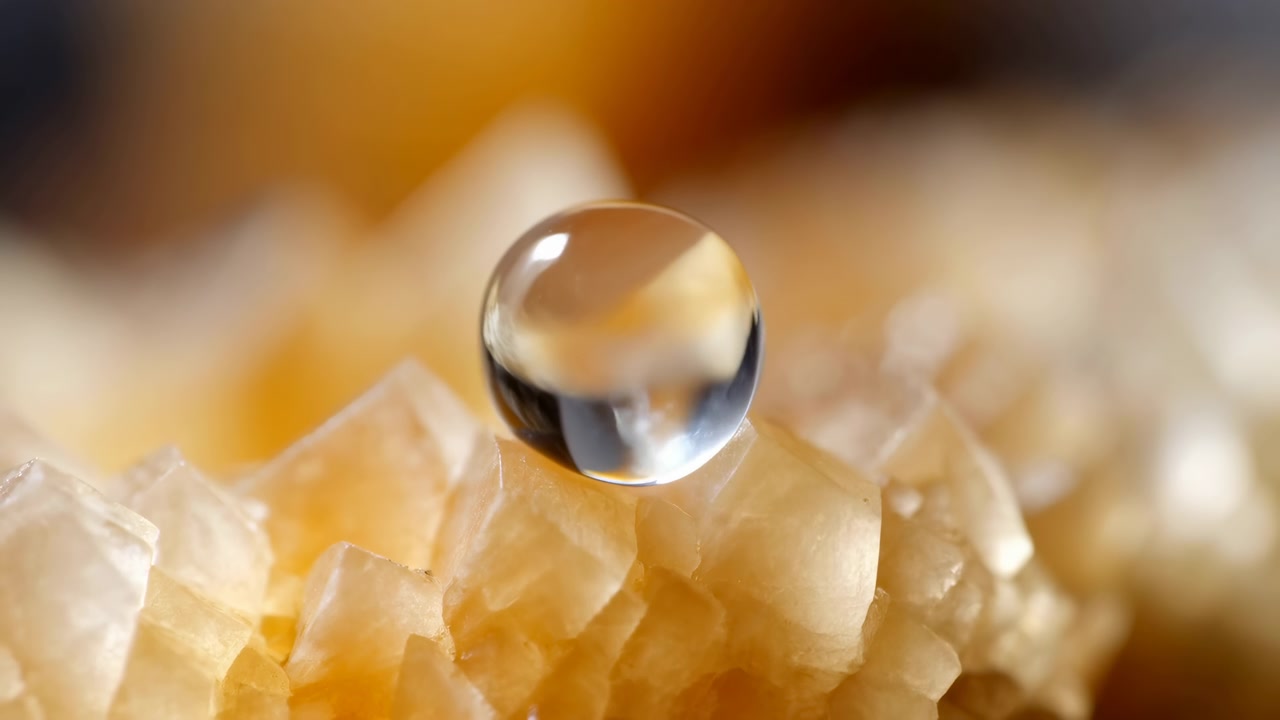 Clear droplet rises above pale amber crystal facets, then returns to gentle contact on textured surface, with warm glow, glossy reflections, soft bokeh, and elegant macro depth, with droplet, water