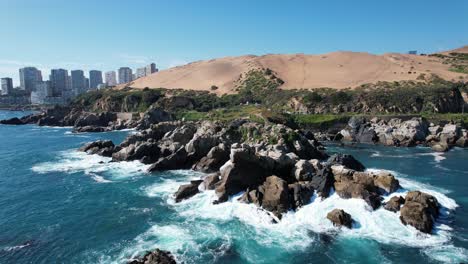 waves breaking on rocks in the coast, with tall buildings and dunes in the background, viña del mar, chile