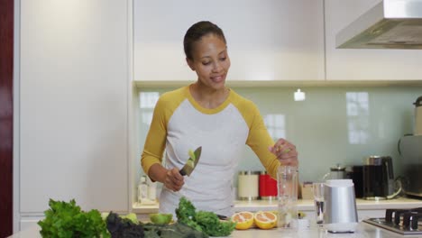 mixed race woman preparing healthy drink, cutting fruit and vegetables in kitchen