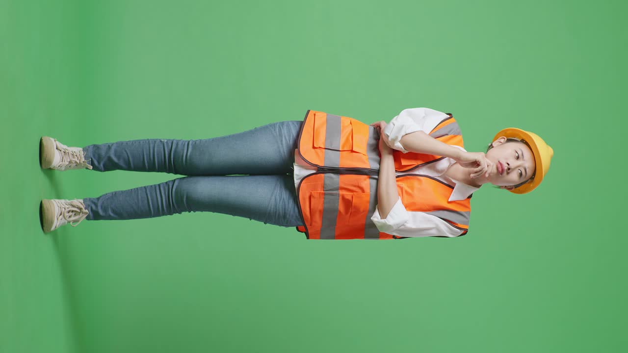 안전 헬을 입은 아시아 여성 엔지니어의 몸이 무언가에 대해 생각하고 녹색 스크린 배경 스튜디오에 서있는 동안 주위를 둘러보고 있습니다.