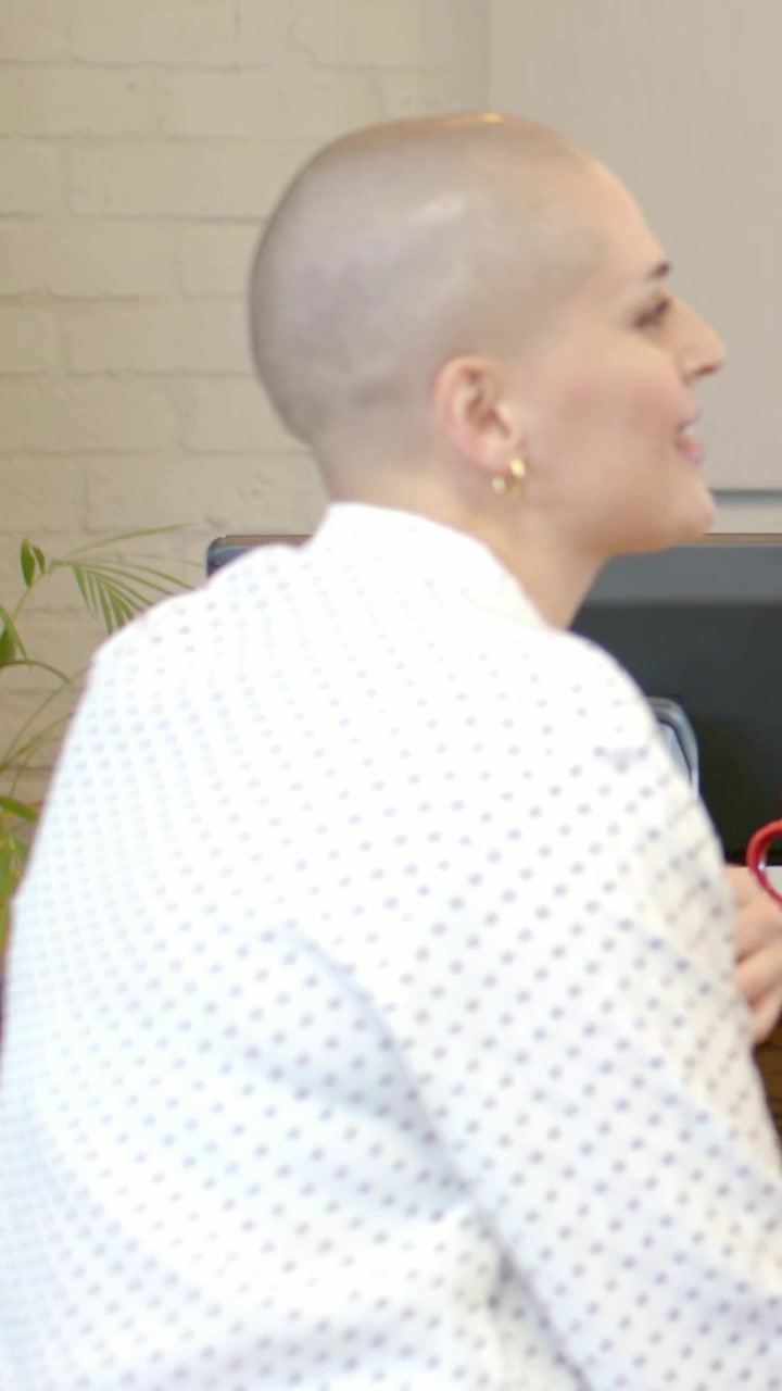 Video verticale: Donna calva in camicia parla alla scrivania, si tocca la bocca e tiene una tazza, sentendo una voce fuori campo.