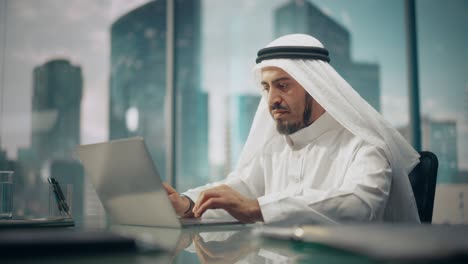 successful arab businessman in white traditional kandura sitting in office and working on laptop computer. business manager planning investments. saudi, emirati, arab businessman concept.