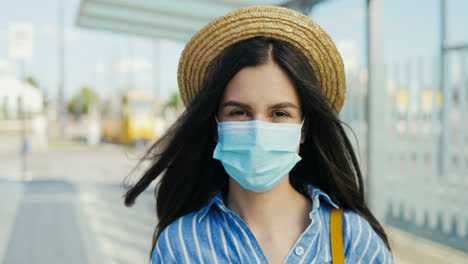 retrato de una joven viajera caucásica con máscara médica y sombrero mirando la cámara al aire libre