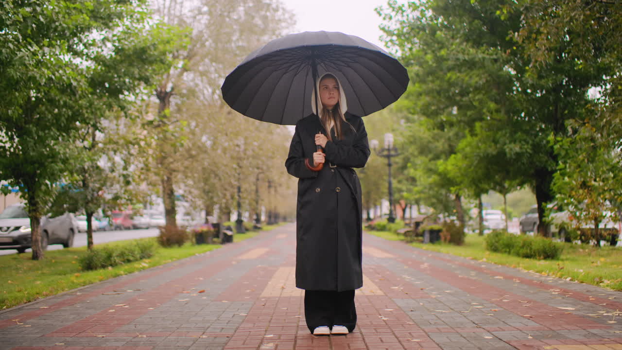 Woman in black trench coat with hood holding umbrella standing on wide city walkway during rainy autumn day, surrounded by trees, benches, parked cars, overcast sky, urban solitude atmosphere