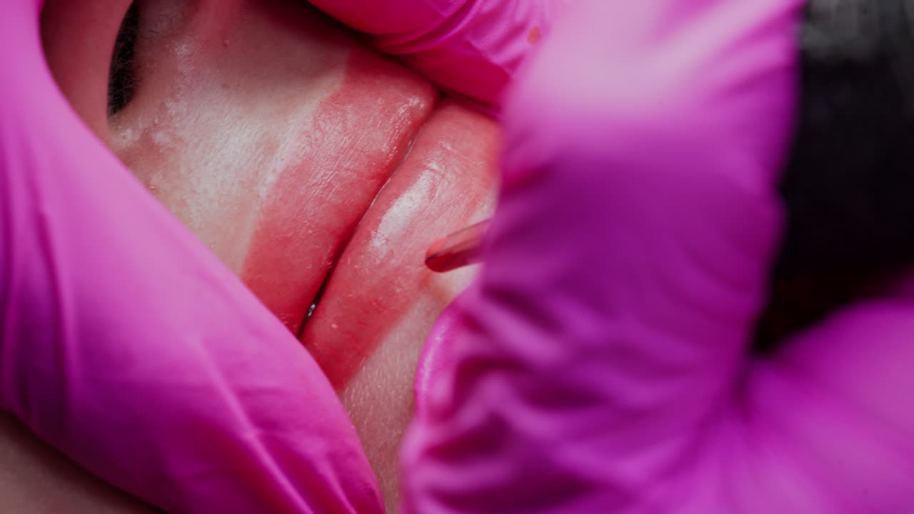Permanent makeup lips procedure. Hands in latex gloves of a skilled cosmetologist applying pigment makeup on lips with a tattoo machine. Macro shot.