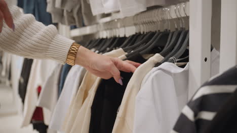 close up female shopper hand with lavender nails, gold watch, chunky knit sleeve shifts hangers, inspecting pastel and striped shirts on store rack, searching right fit during calm retail browse