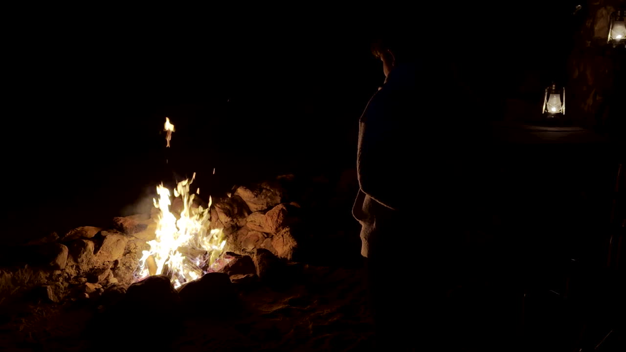 Teenage boy mesmerized by African campfire flames, paraffin lamps in background