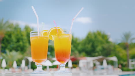 two orange cocktails with straws and orange slices are on the beach table by the pool against the bl