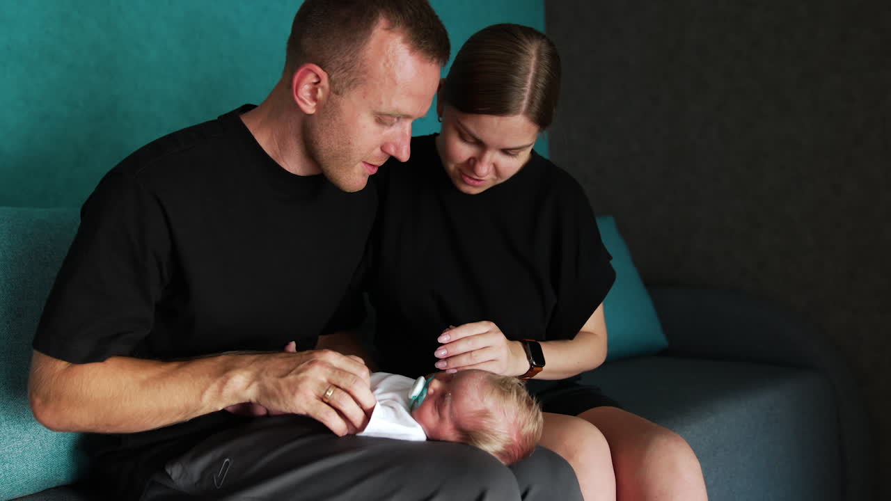 New-made parents admiring their cute newborn baby. Kid lies on dad's laps sleeping and parents touch his tiny hands and nose.