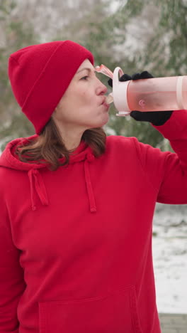 lady in red hoodie and beanie sipping water from pink bottle outdoors in winter park with snowy ground, green trees, and bench in background, peaceful winter setting