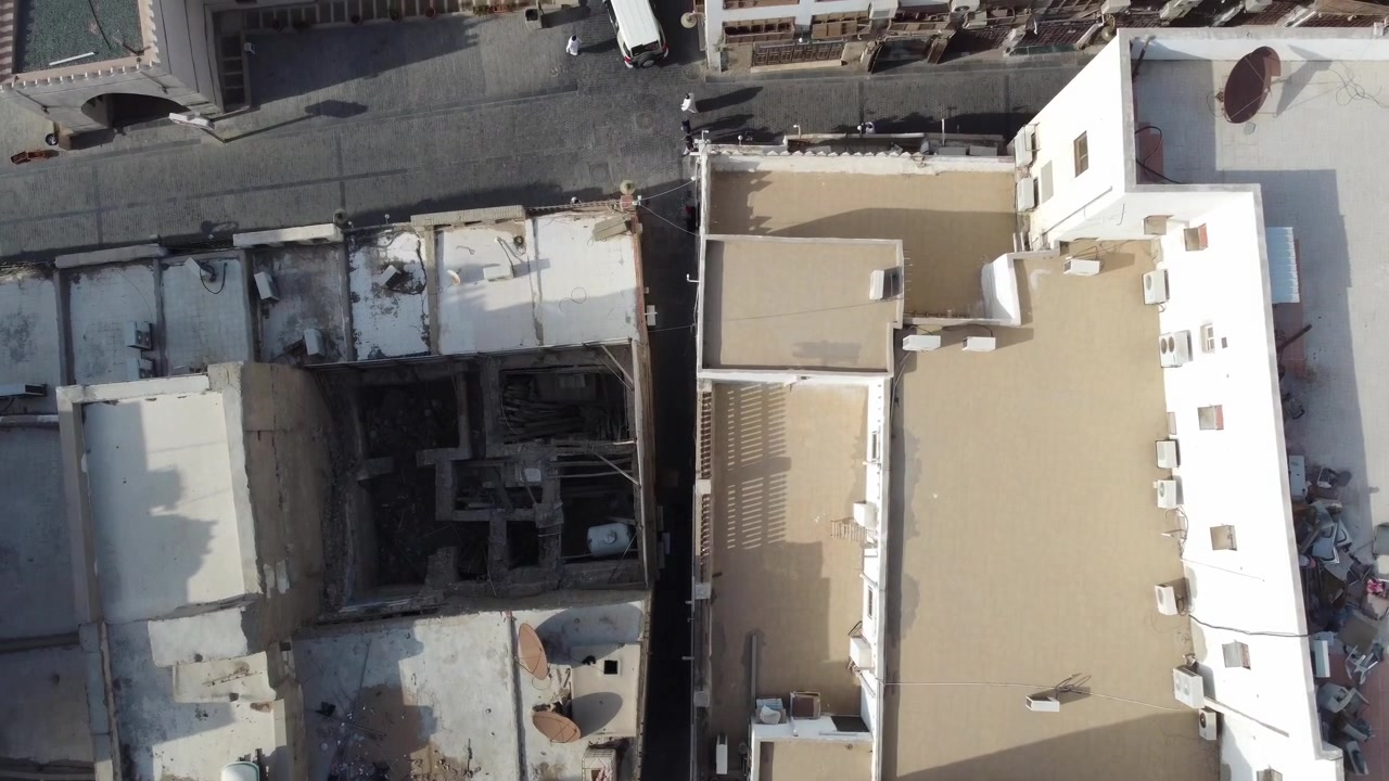 Ripresa aerea zenitale diretta su fitti tetti urbani, antenne paraboliche, unità di condizionamento e un vicolo stretto in un vecchio quartiere residenziale dell'Arabia Saudita sotto la luce solare intensa
