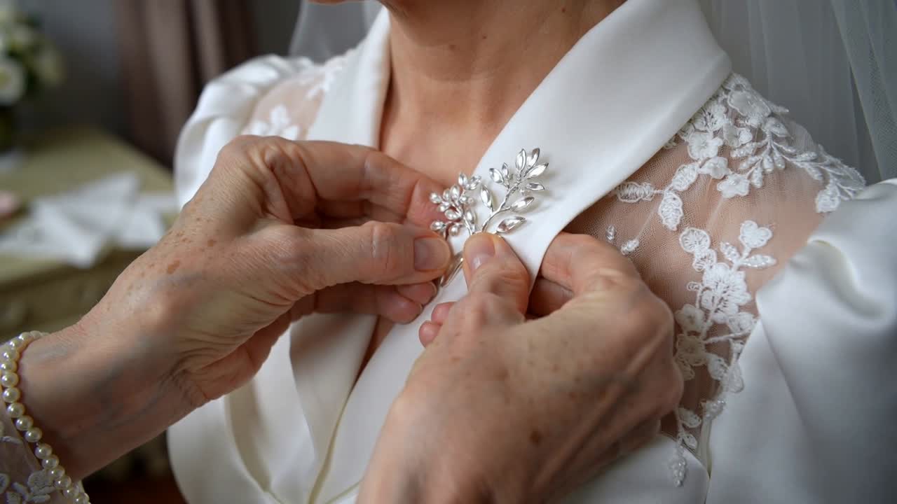 Bringing silver leaf brooch, assistant fastening pin on woman in white gown in bedroom, for wedding
