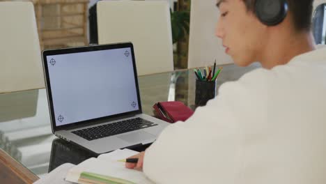 Asian-male-teenager-with-headphones-learning-and-using-laptop-in-living-room