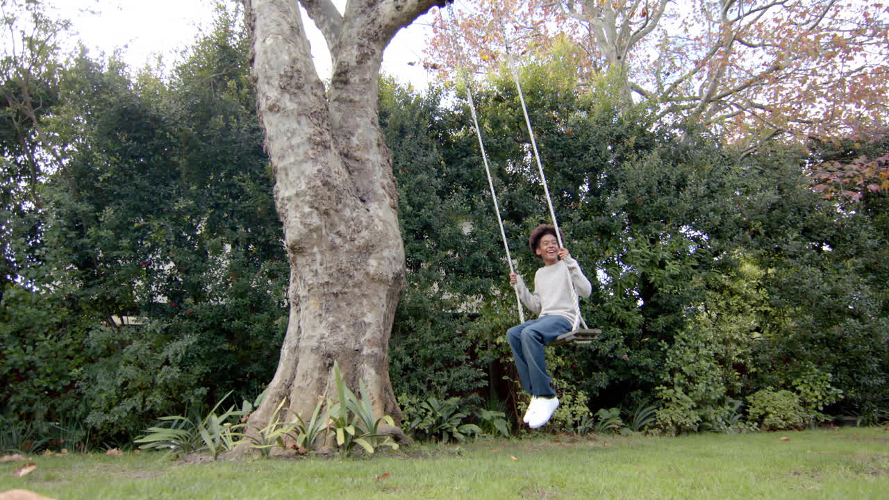 Happy african american boy swinging on tree swing in garden, slow motion