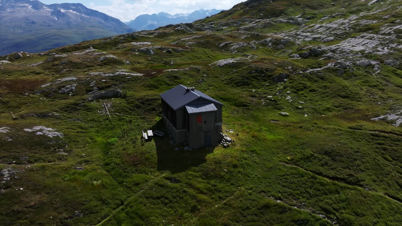 Aerial view of solitary cabin rests on a verdant hillside, surrounded by rocky terrain under a clear sky, evoking a sense of isolation and tranquility in the wilderness.