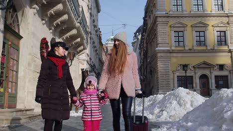 two young smiling women tourists with adoption child girl walking with suitcase on old city streets