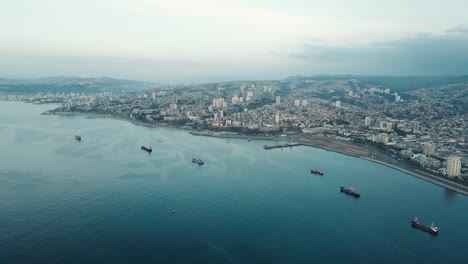 Aerial-parallax-of-Valparaiso-hillside-city-and-Sea-Port-with-container-cargo-ship-sailing-near-the-coastline,-Chile