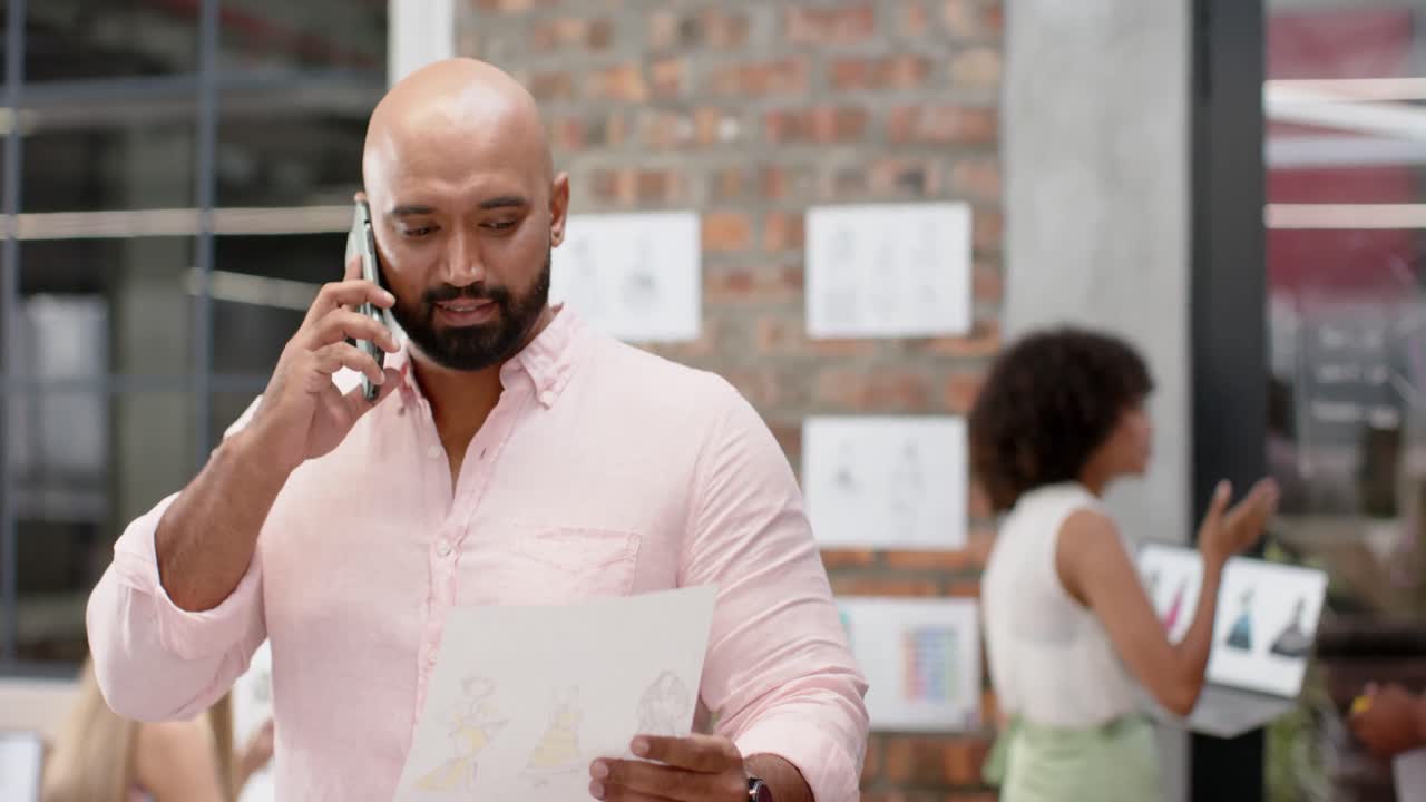 Happy biracial male creative holding document and talking on phone at fashion studio, in slow motion