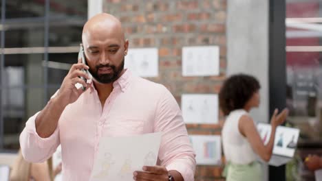 happy biracial male creative holding document and talking on phone at fashion studio, in slow motion