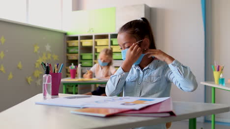 Kleines-Mädchen-Setzt-Im-Unterricht-Eine-Medizinische-Maske-Auf.-Covid-19-Pandemie.-Kinder-Gehen-Nach-Der-Sperrung-Des-Coronavirus-Wieder-Zur-Schule.