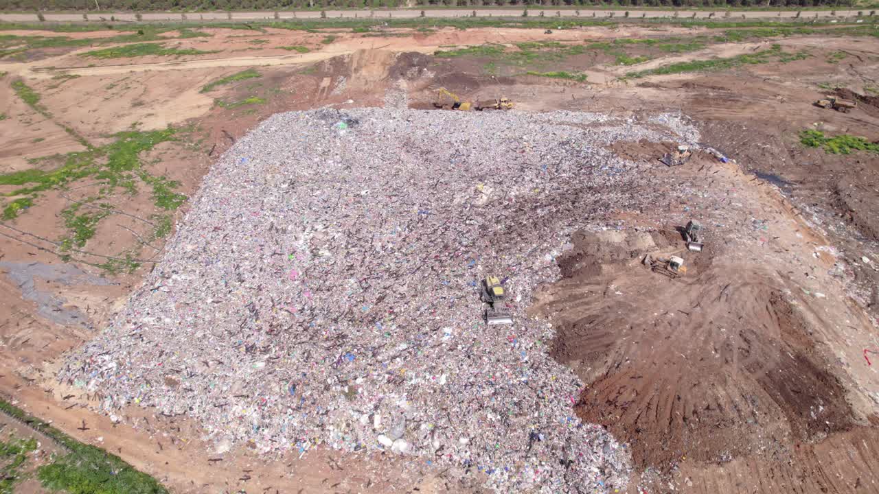 Aerial drone shot of a bulldozer operating on a large landfill mound while dozens of birds of prey circle and fly above the waste site, highlighting human impact, waste management activity, and wildli